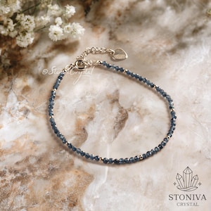 Puede incluir: Delicada pulsera con pequeñas gemas azules facetadas y cuentas doradas. La pulsera tiene una cadena plateada con un dije en forma de corazón. El fondo es una superficie similar al mármol con flores blancas. La marca "STONIVA CRYSTAL" es visible.