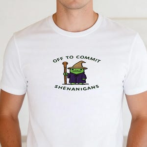 Może przedstawiać: Biała koszulka z postacią z kreskówki żaby w stroju czarodzieja, trzymającej laskę. Tekst "OFF TO COMMIT SHENANIGANS" jest wydrukowany nad i pod postacią. Projekt jest zielony, czarny i brązowy.
