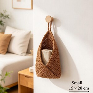 May include: A small, brown, crocheted hanging basket with a braided handle, suspended from a round wooden wall hook. The basket contains a folded beige cloth and is labeled with the dimensions 15 x 28 cm in the bottom corner.