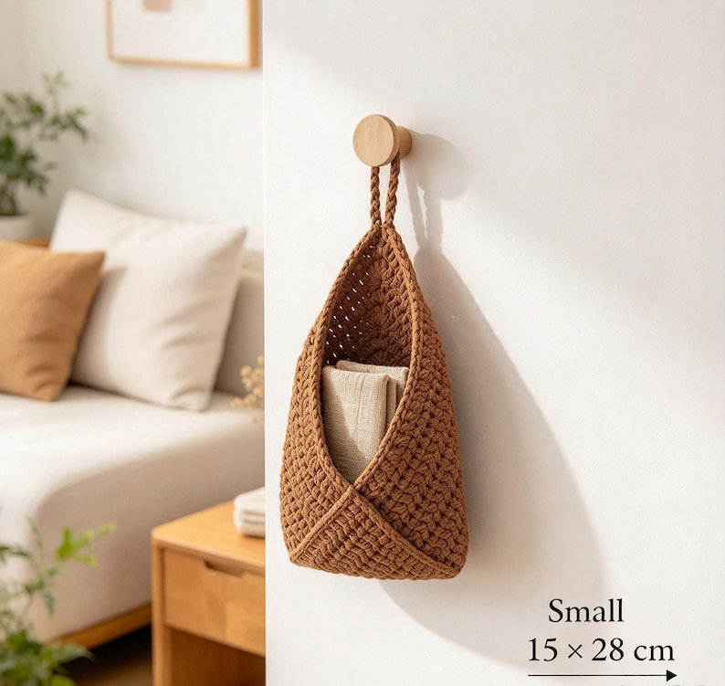 May include: A small, brown, crocheted hanging basket with a braided handle, suspended from a round wooden wall hook. The basket contains a folded beige cloth and is labeled with the dimensions 15 x 28 cm in the bottom corner.