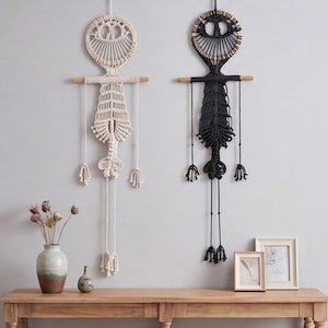 May include: Two macrame wall hangings, one in cream and one in black, each featuring a circular top, a horizontal wooden dowel, and a long, intricate knotted body with dangling fringe elements, suspended vertically against a plain wall.