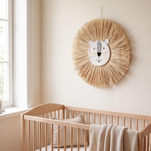 May include: A circular wall hanging featuring a stylized lion face made of white coiled rope with a grey nose and black embroidered eyes and mouth. The lion has a large, radiating mane made of natural tan raffia or straw-like fibers.
