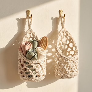 May include: Two off-white macrame hanging baskets suspended from brass-coloured wall hooks. The left basket contains three rolled silk cloths in shades of dusty pink, sage green, and cream, alongside a wooden hairbrush with rounded bristles.