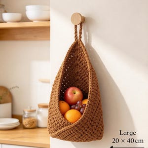 May include: A brown crocheted hanging basket with a braided handle suspended from a wooden wall hook. The basket contains an apple, two oranges, and a cluster of grapes. Text on the image indicates a size of 20 x 40 cm.