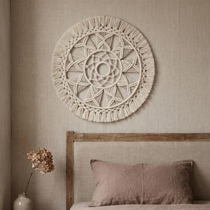 Puede incluir: Un tapiz de pared de macramé color crema con un diseño circular. El intrincado patrón presenta formas geométricas y un borde con flecos. Esta pieza decorativa añade un toque bohemio a cualquier habitación. El diámetro es de aproximadamente 61 cm.