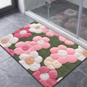 May include: A rectangular bath mat featuring a repeating floral pattern with large, plush, tufted flowers in shades of pink, white, and beige against a dark olive green background. The mat has a soft, textured surface and a clean, finished edge.