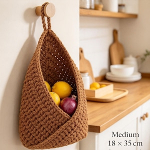 May include: A medium-sized brown crocheted hanging basket measuring 18 x 35 cm, suspended from a wooden wall hook. The basket features a thick, textured knit pattern and contains three yellow lemons and one red onion inside its open, tapered pocket.
