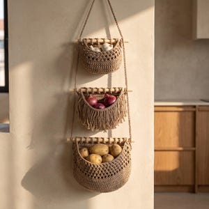 May include: A three-tiered hanging storage organizer made of tan crocheted cord suspended by wooden dowels. The top basket holds garlic, the middle basket with fringe holds red onions, and the bottom basket holds several brown potatoes.
