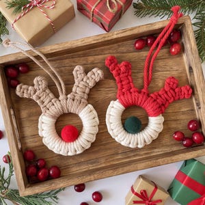 May include: Two macrame reindeer ornaments featuring circular bases and knotted antlers. One ornament is beige with a red pom-pom nose, and the other is red with a dark green pom-pom nose. Both include hanging cords for display as holiday decorations.