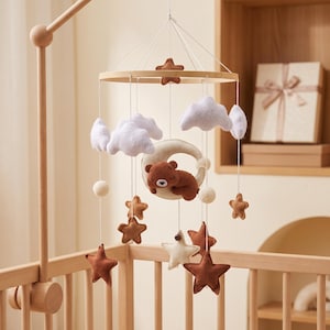 May include: A nursery mobile featuring a circular wooden frame suspended with white strings. It includes felt elements: a brown bear sleeping on a cream crescent moon, white clouds, and various stars in shades of brown, cream, and tan.