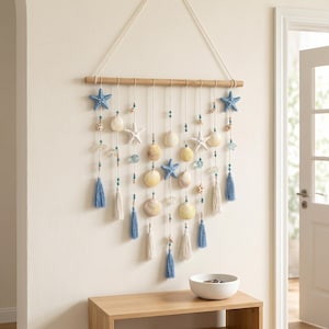 May include: A wall hanging featuring a horizontal wooden rod suspended by a cord. Multiple vertical strands hang from the rod, decorated with blue and white starfish, various seashells, small blue and tan beads, and blue and white fabric tassels.