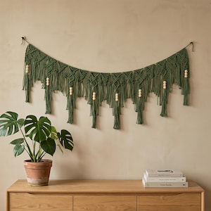 May include: A wall hanging made of olive green macrame cord featuring intricate knotted patterns and dangling fringe. The piece is accented with light-colored wooden beads integrated into the design and is suspended from two small metal hooks.