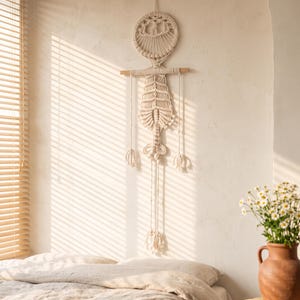 May include: A cream-colored macrame wall hanging featuring a circular top section with intricate knotting, suspended from a horizontal wooden dowel. The design includes a central leaf-like pattern with long, dangling cords ending in small tassels.