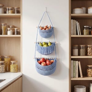 May include: A three-tiered hanging storage organizer made of light blue crocheted fabric and wooden dowels. The top basket holds onions, the middle basket contains green pears with fringe detailing, and the bottom basket is filled with red apples.