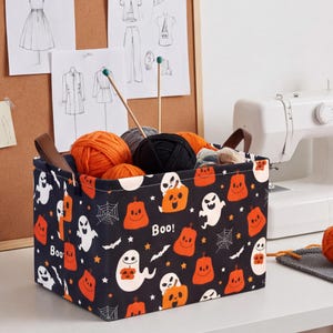 Puede incluir: Una cesta de almacenamiento rectangular de tela negra con un patrón repetitivo de Halloween de calabazas naranjas, fantasmas blancos, telarañas y el texto "Boo!". La cesta tiene asas de piel sintética marrón y está llena de ovillos de lana.