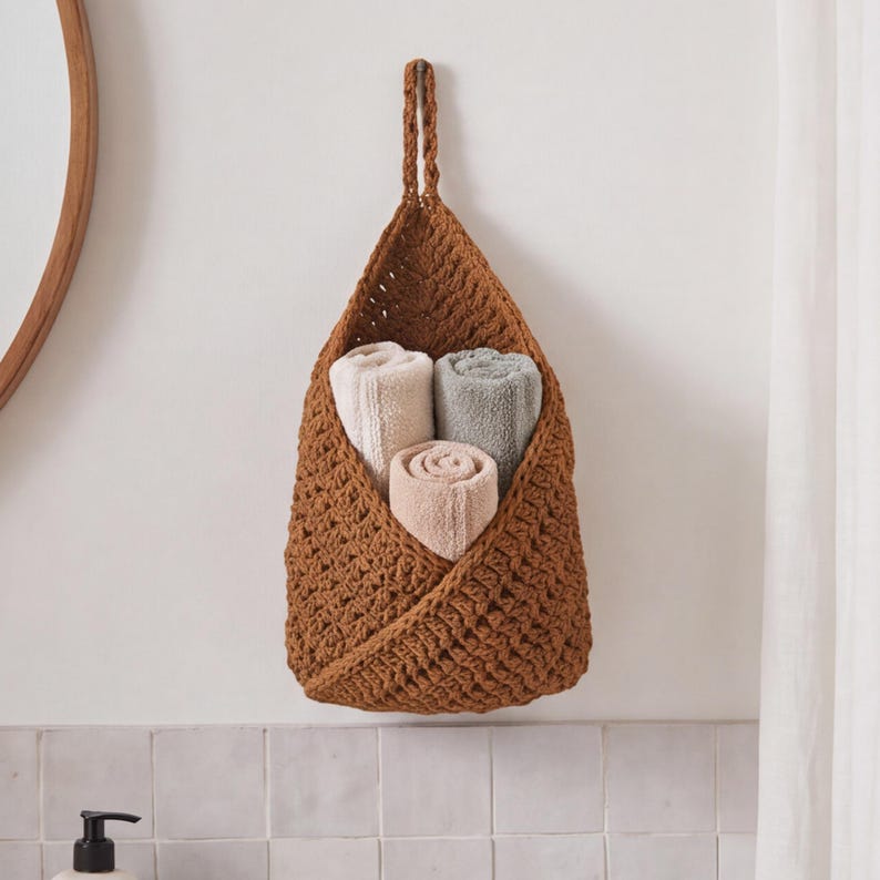 May include: A hanging storage basket made of thick, brown crocheted yarn with a triangular shape and a top loop. Inside the basket are three rolled towels in shades of cream, light gray, and soft pink, neatly stacked for storage.