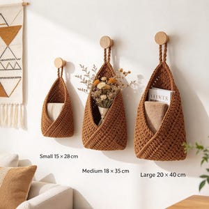 May include: Three brown crocheted hanging baskets with braided loops suspended from round wooden wall hooks. The baskets are labeled with dimensions: small 15 x 28 cm, medium 18 x 35 cm, and large 20 x 40 cm. They contain dried flowers and folded fabric.