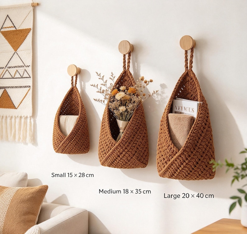 May include: Three brown crocheted hanging baskets with braided loops suspended from round wooden wall hooks. The baskets are labeled with dimensions: small 15 x 28 cm, medium 18 x 35 cm, and large 20 x 40 cm. They contain dried flowers and folded fabric.