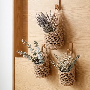 May include: Three tan, hand-crocheted hanging baskets made of thick cord, each featuring an open-weave diamond pattern. The baskets are suspended from wooden wall hooks and contain dried lavender, eucalyptus stems, and white baby's breath flowers.
