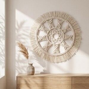 May include: A circular off-white macrame wall hanging featuring a central woven mandala pattern, radiating geometric spokes, and a thick outer fringe. The piece is constructed from knotted cotton cord, creating a textured, symmetrical design.