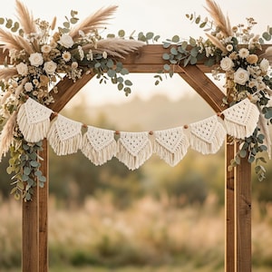 Könnte beinhalten: Eine hölzerne Hochzeitsbogen mit weißen Blumen, Pampasgras und Grünzeug geschmückt. Ein Makramee-Banner mit dreieckigen Anhängern hängt über dem Bogen. Die Szene spielt sich im Freien mit einem verschwommenen Feld im Hintergrund ab.