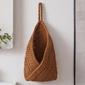 May include: A hanging storage basket crafted from brown yarn with a textured crochet stitch pattern. The basket features a deep, open-front design with a braided loop at the top for suspension, providing a functional storage solution.