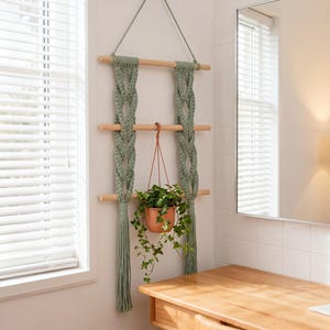 May include: A wall-hanging macrame plant shelf featuring three horizontal light-colored wooden dowels connected by sage green knotted cord. A terracotta-colored pot containing a green ivy plant hangs from the center dowel by a cord.