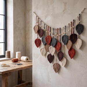 May include: A macrame wall hanging featuring multiple leaf-shaped fiber ornaments in shades of cream, tan, rust, dark red, brown, and charcoal gray, suspended from a central cord with small wooden beads interspersed between each hanging element.