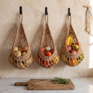 May include: Three beige woven jute hanging baskets suspended from black wall hooks. The left basket holds potatoes, the center basket contains tomatoes and garlic, and the right basket stores bananas, apples, and oranges. A wooden cutting board sits below.