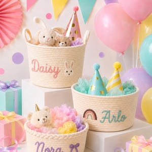 May include: Three off-white woven rope baskets featuring embroidered names and designs. One basket displays the name Daisy with a rabbit face, another shows Arlo with a rainbow, and the third features Nora with a bow. Each contains plush toys and party hats.