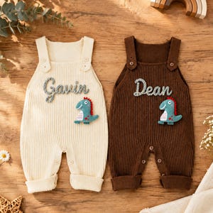 May include: Two ribbed knit rompers, one cream and one dark brown, each featuring shoulder buttons, a cuffed hem, and a button-up crotch. The cream romper displays the name Gavin in cursive, and the brown romper displays the name Dean, both with a small teal dinosaur appliqué.