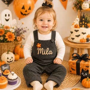 May include: A dark grey ribbed knit overall with a white long-sleeve shirt underneath. The overall features the embroidered name Mila in a light-coloured script font and a small orange flower appliqué on the chest, secured with dark buttons.