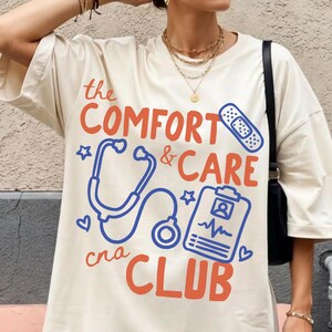 May include: Ivory-colored t-shirt with the text "The Comfort & Care Club" in orange and blue. The design includes a stethoscope, a bandage, and a medical chart. The model is wearing red and white checkered shorts.