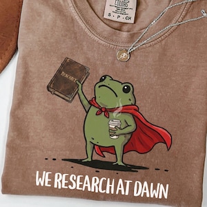 May include: A brown short-sleeve t-shirt featuring a graphic of a green frog wearing a red cape, holding a brown book labeled "RESEARCH" and a white coffee cup. The text "WE RESEARCH AT DAWN" is printed in white below the frog illustration.