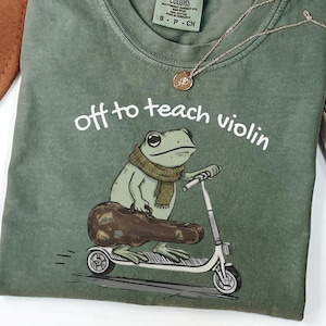 May include: Moss green t-shirt with the text "off to teach violin." The graphic shows a frog wearing a scarf, riding a scooter, and carrying a violin case. A gold necklace and a brown hat are also visible.