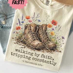 May include: An ivory t-shirt featuring a graphic of two brown hiking boots surrounded by colorful wildflowers. The text reads, "walking by faith, tripping constantly" followed by "-2 Corinthians 5:7a-" in a black typewriter-style font.
