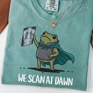 May include: A light green t-shirt featuring a graphic of a frog wearing a grey chest plate and a teal cape, holding up an X-ray image. Below the frog, the text reads "WE SCAN AT DAWN" in white capital letters. The shirt has a Comfort Colors label.