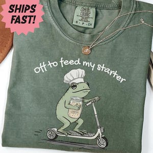 May include: Moss green t-shirt with the phrase "off to feed my starter" and a cartoon frog chef riding a scooter. The frog is holding a jar. A pink starburst sticker says "SHIPS FAST!"