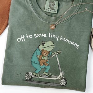 May include: Moss green t-shirt with the phrase "off to save tiny humans" in white. The graphic features a frog in scrubs riding a scooter and holding a teddy bear. A gold necklace is draped across the shirt.