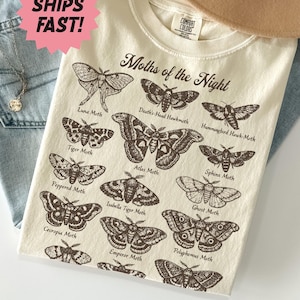May include: Ivory-colored t-shirt featuring a detailed illustration of various moth species, including Luna Moth and Atlas Moth, with the text "Moths of the Night." The shirt is folded on top of blue jeans.