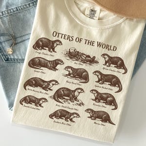 May include: An ivory t-shirt featuring a brown graphic print titled Otters of the World, displaying illustrations of various otter species including the Sea Otter, Giant Otter, and Marine Otter, with each species clearly labeled in brown text.