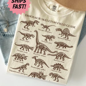 May include: An ivory t-shirt featuring a brown graphic print titled Dinosaur Skeleton Field Guide, displaying various dinosaur skeletons including Tyrannosaurus, Velociraptor, Spinosaurus, Allosaurus, Carnotaurus, Ankylosaurus, Diplodocus, Brachiosaurus, Apatosaurus, Triceratops, Parasaurolophus, Stegosaurus, Pachycephalosaurus, and others.