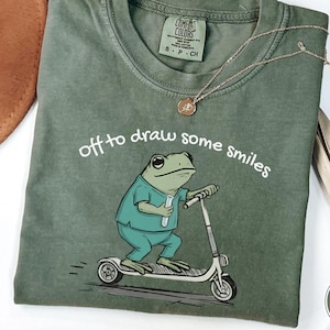 May include: Moss green t-shirt with a frog graphic riding a scooter and the text "off to draw some smiles." The frog is wearing a teal uniform and holding a vial. A gold necklace and a brown hat are also visible.