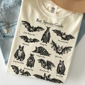 May include: Ivory-colored t-shirt featuring a "Bat Species Guide" graphic with various bat illustrations and names. The shirt is laid on top of blue jeans and a tan hat. The text "Ivory" is at the bottom left.