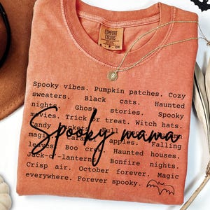 May include: A burnt orange short-sleeved t-shirt featuring black text listing Halloween-themed phrases like Spooky vibes, Pumpkin patches, and Haunted houses, with the script text Spooky mama across the center and a small bat graphic at the bottom.
