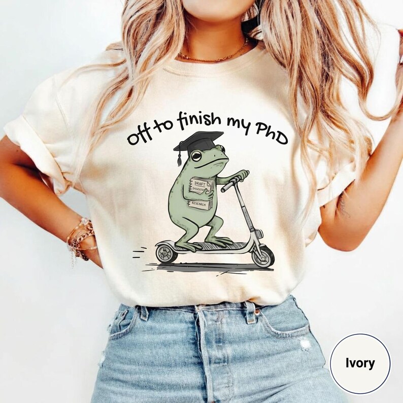 May include: Ivory-colored t-shirt with the text "off to finish my PhD." The graphic features a cartoon frog wearing a graduation cap and riding a scooter while holding a book labeled "Draft Research."