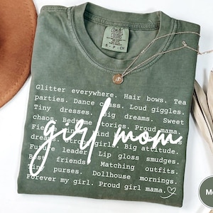 May include: A moss green t-shirt with the words "girl mom" in a cursive font. The shirt features a list of words related to motherhood, such as "glitter," "parties," and "big dreams." A gold necklace with a pendant rests on the shirt.