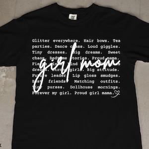 May include: Black t-shirt with white text. The text includes phrases like "girl mom," "glitter everywhere," and "proud girl mama." A heart symbol is at the end of the text. The shirt is a casual style.