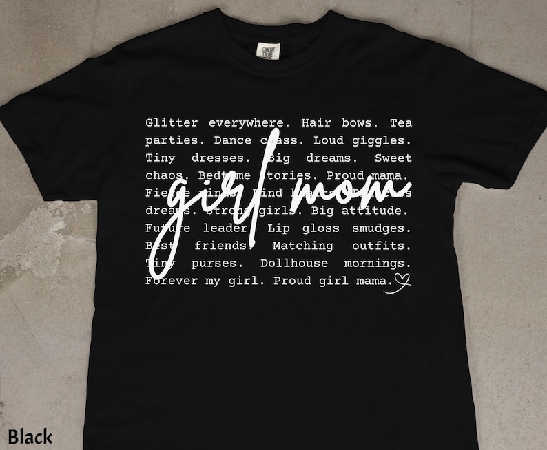 May include: Black t-shirt with white text. The text includes phrases like "girl mom," "glitter everywhere," and "proud girl mama." A heart symbol is at the end of the text. The shirt is a casual style.