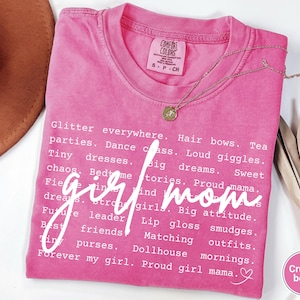 May include: A pink Comfort Colors t-shirt with the words "girl mom" in white script. The shirt features a list of related words like "glitter," "dreams," and "proud mama." A gold necklace with a pendant is draped on the shirt.